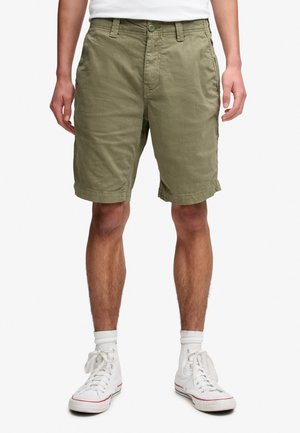 Mand iført olivengrønne knælange shorts, hvide high-top sneakers og hvide sokker, står mod en ensfarvet hvid baggrund.