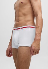 Boxers blancs avec une texture douce, présentant une ceinture élastique rouge avec le logo "HUGO". Le design est ajusté et simple, sans motifs.