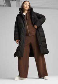 Doudoune noire avec capuche, associée à un sweat à capuche en polaire marron et un pantalon large. Des baskets blanches complètent la tenue.