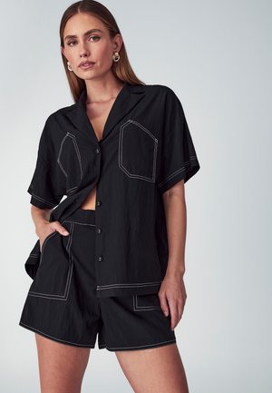 Schwarzes Shirt- und Shorts-Set aus strukturiertem Stoff. Das Shirt hat kurze Ärmel, eine Knopfleiste und zwei kontrastierend genähte Taschen.