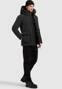 Schwarze gepolsterte Jacke mit Kapuze, zwei Fronttaschen und zentralem Reißverschluss. Wird über einem schwarzen Pullover getragen und mit schwarzen Hosen und Stiefeln kombiniert.