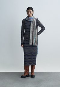 Hosszú, testhez simuló ruha geometriai mintával tengerészkék és szürke színekben. Egy több színű cikk-cakk sállal és barna térdig érő csizmával párosítva.