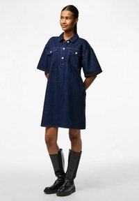 Tumši zila džinsu kleita ar apkakli, divām krūšu kabatām, īsām piedurknēm un pogu aizdari. Valkāta ar līdz ceļgaliem melnām zābakiem.