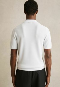 Weißes kurzärmliges Poloshirt aus strukturiertem, geripptem Stoff mit einem flachen Kragen. Das Shirt ist mit schwarzen Hosen kombiniert und von hinten betrachtet.