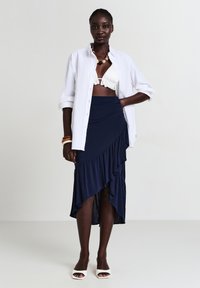 Donna che indossa una camicia bianca aperta sopra un top bikini bianco con volant, una gonna asimmetrica navy con volant, tacchi open-toe bianchi e braccialetti voluminose.