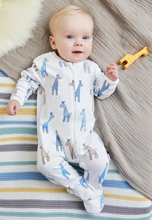 Smiling baby in white footed pajamas with blue and beige giraffe prints lying on striped knitted blanket and gray quilt.