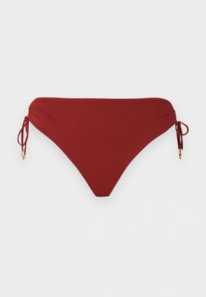 Kobieta ubrana w biały top od bikini, brązowe majtki bikini i czarne klapki, stojąca z jedną ręką na biodrze na jednolitym tle.