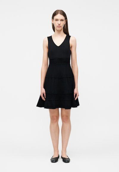 Femme debout, face à l'avant, portant une robe noire sans manches avec des bandes horizontales texturées et des chaussures plates noires, devant un fond uni.