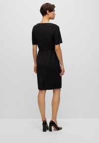Black short-sleeve dress with a fitted silhouette, back zip closure, and a subtle waist tie. Smooth fabric with a knee-length hem.