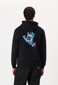 Melnā kapuce ar lielu, zilu zīmējumu, kurā attēlota roka un mute, uz muguras. Mīksta auduma, regulējama kapuce, ribota aproce un apakšmala.