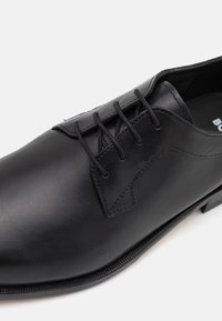 Black leather shoe with round toe, lacing system, subtle stitching details, and a flat sole. Smooth texture, minimalistic design.