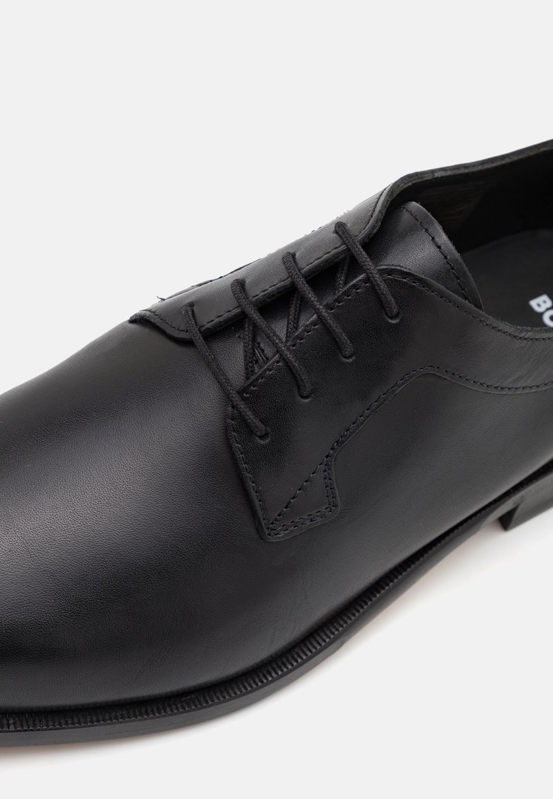 Chaussure en cuir noir à bout rond, système de laçage, détails de couture subtils et semelle plate. Texture lisse, design minimaliste.