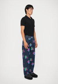 Tricou polo negru asortat cu pantaloni din denim albastri, cu modele colorate de pahare de băutură în stil cartoon, și pantofi slip-on negri.