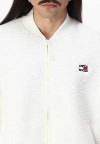 Man met snor die een wit ritsjack van sherpa draagt met een klein Tommy Jeans-logo op de borst.