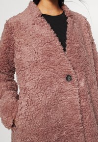 Lyserød kunstpelsjakke med en tekstureret, fluffy overflade. Har sort knaplukning og brede revers. Designet inkluderer sidelommer.