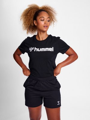 Жена с къдрави коси, облечена в черна тениска hummel и къси панталони, коригира късите си панталони, докато гледа настрани на бял фон.