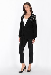Czarny kardigan z wiązaniem w talii, z perłowymi akcentami na ramionach. Noszony na białej bluzce, w zestawieniu z czarnymi spodniami i szpilkami.
