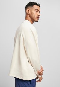 Pulover bej oversized cu guler rotund, umeri lăsați și margini ribbate, realizat dintr-un material moale, expus alături de pantaloni închisi la culoare.