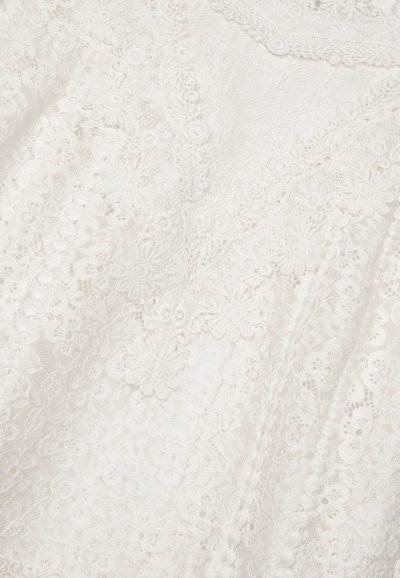 Tissu en dentelle blanche présentant des motifs floraux, des détails raffinés, des bords festonnés et une finition texturée, créant une apparence délicate.