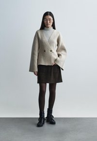 Beige dubbelknäppt jacka i ull med klockärmar, kombinerad med en randig polotröja, mörk mockakjol och svarta snörstövletter.