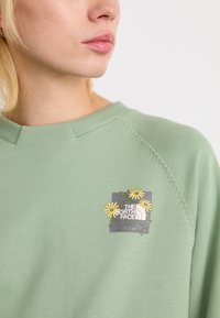 Sweatshirt vert avec un col rond, arborant un logo floral avec des fleurs jaunes et la marque The North Face sur la partie gauche de la poitrine.