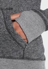 Grå textilhoddie med känguruficka och kontrasterande ribbad mudd. Materialet har en mjuk, texturerad yta med synliga sömnadsdetaljer.