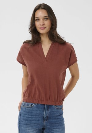 Vrouw met lang bruin haar, gekleed in een roestkleurige top met korte mouwen en een V-hals, en een blauwe spijkerbroek, staand met handen in de zakken.