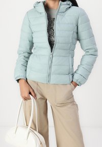 Lyseblå pufferjakke med hætte, med quiltet tekstur, frontlynlås og elastiske manchetter, båret over en mønstret top og beige bukser.