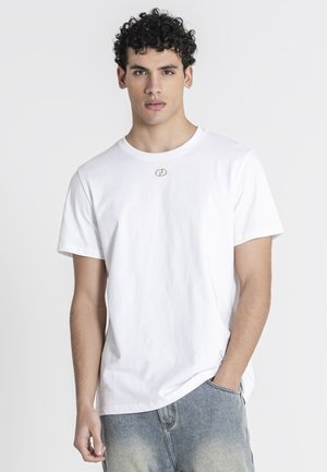 Giovane uomo con capelli ricci che indossa una t-shirt bianca semplice a maniche corte e jeans azzurri, in piedi davanti a uno sfondo chiaro uniforme.