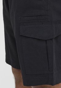 Mustat cargo-shorts, jotka on valmistettu teksturoidusta kankaasta ja joissa on sivutasku, jossa on läppä. Ompeleet näkyvät helman ja saumoissa.