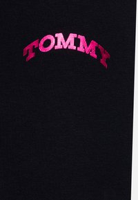 Crni materijal s rebrastom teksturom s ružičastim izvezenim logotipom "TOMMY" u luku na gornjem dijelu.