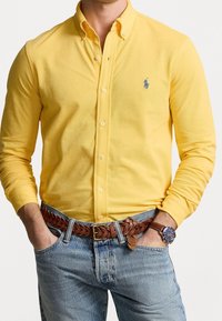 Żółta koszula z długim rękawem, zapinana na guziki, z kołnierzykiem i akcentem w postaci logo, zestawiona z niebieskimi dżinsami i plecionym brązowym skórzanym paskiem.