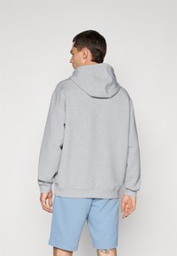 Mand, der står med ryggen til, iført en lysegrå hættetrøje med huen oppe og lyseblå shorts, stillede op mod en ensfarvet lys baggrund.