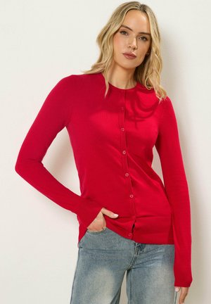 Frau mit blondem Haar, die eine leuchtend rote, geknöpfte Strickjacke und hellblaue Jeans trägt, steht vor einer schlichten weißen Wand.