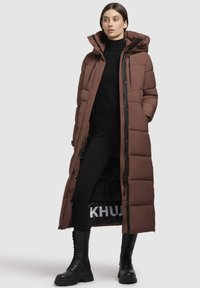 Ruda pūkuota striukė su kapišonu, ilgai dizainu, užtrauktuku ir siuvinėtu tekstūru. Dėvima ant juodos aprangos ir tvirtų juodų batų.