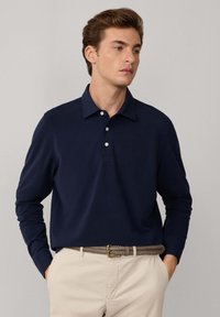 Granatowa koszula polo z długim rękawem, z kołnierzykiem i zapięciem na trzy guziki, w połączeniu z jasnobeżowymi spodniami i plecionym paskiem.
