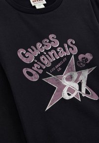 Schwarzes Baumwoll-T-Shirt mit einem abgenutzten Grafikdesign in Hellpurpur und Silber, das "Guess Originals" und ein Sternenmuster mit der Zahl "81" zeigt.