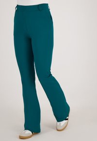 Tegengeschiedenis bootcut broek in de kleur teal, gedragen met witte sneakers met zwarte strepen en rubberen zolen op een lichte effen achtergrond.