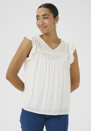 Vrouw met donker haar in een staart, gekleed in een mouwloos wit blouse met gerimpelde schouders en een blauwe spijkerbroek, licht glimlachend opzij.