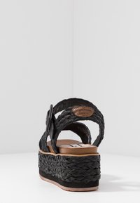 Svart vävd plattformssandal med flätade remmar, läderdetalj och en brun dämpad sula, med en lätt klack och texturerad yta.
