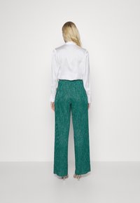 Bílá blůza s dlouhým rukávem doplněná širokými kalhotami v barvě teal s texturovaným vzorem. Outfit doplňují průhledné podpatky.