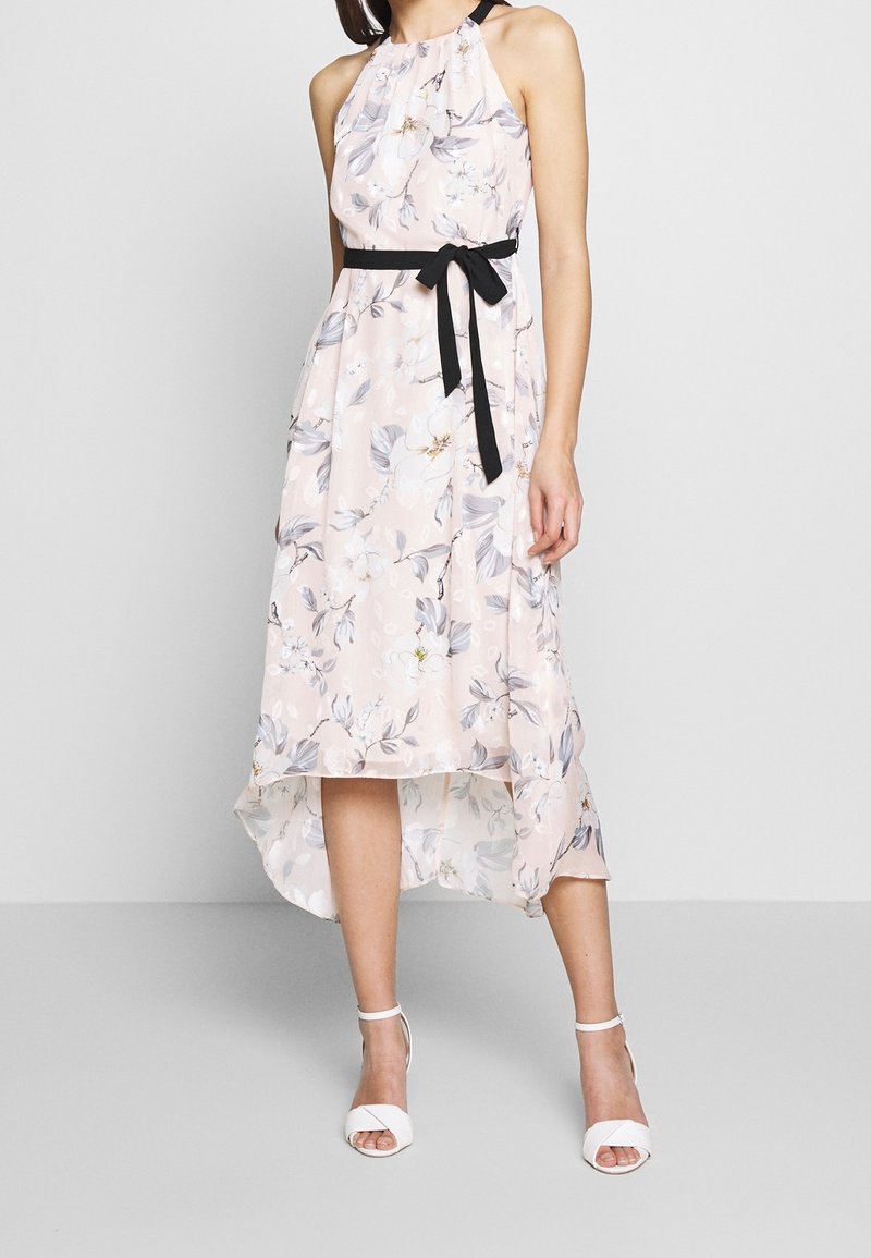 Femme portant une robe sans manches rose pâle à fleurs avec un ourlet asymétrique et une ceinture noire à la taille, associée à des talons blancs à bride cheville.