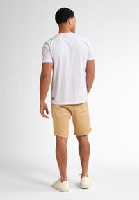 Hvid kortærmet t-shirt, løs pasform, parret med tan cargo shorts. Casual sneakers fuldender outfittet. Bagview med ensfarvet baggrund.