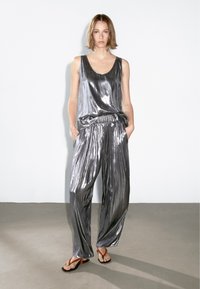 Metallic silver ärmlös topp som bärs med plisserade silver vida byxor. Accenterad av ett elastiskt midjeband, bärs med enkla sandaler.