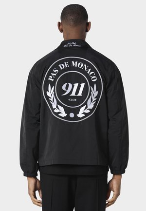 Mand iført sort jakke med hvidt emblem "PAS DE MONACO 911 CLUB" og kransingdesign på ryggen, stående mod en lys, ensartet baggrund.