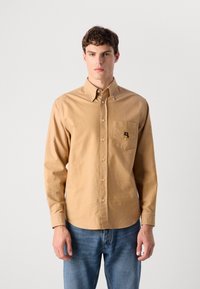 Mann iført en beige knapp-up skjorte med brystlomme og brodert logo, kombinert med blå jeans, stående mot en ensfarget bakgrunn.
