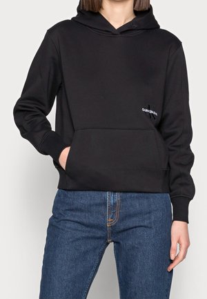 Osoba nosząca czarną bluzę z kapturem Calvin Klein z przednią kieszenią i niebieskie dżinsy, ręka w kieszeni, stojąca na jednolitym tle.