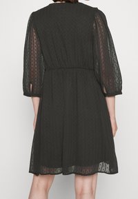 Svart klänning i lätt tyg med genomskinliga puffärmar och prickig textur; har en åtsittande midja och en utsvängd nederkant.