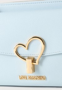 Γαλάζια δερμάτινη τσάντα με χρυσό κούμπωμα σε σχήμα καρδιάς και το λογότυπο "LOVE MOSCHINO" με χρυσά γράμματα κάτω από το κούμπωμα.
