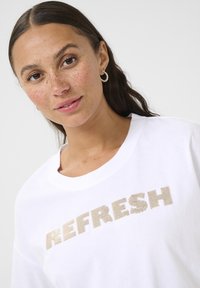 T-shirt en coton blanc avec "REFRESH" en lettres dorées texturées. Le modèle a de longs cheveux et porte des boucles d'oreilles créoles, affichant un teint naturel.